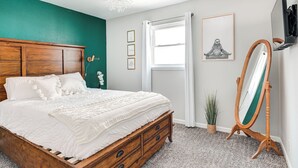 Une chambre avec un lit en bois, un mur d’accent vert, une fenêtre avec des rideaux, un tableau encadré, un miroir et une plante en pot.