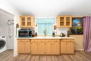 A compact kitchen with wooden cabinets, a microwave, a coffee maker, and a window with a view of a landscape. - 3-bedroom house , Elvia’s country style . (Perris)