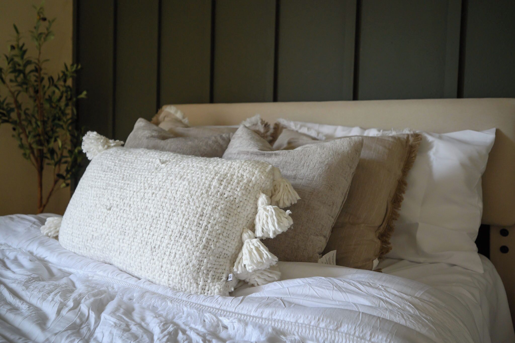 Una cama con ropa de cama blanca y varios almohadones, incluyendo uno grande de color blanco con borlones.