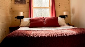 Una habitación con paneles de madera, una cama, dos mesitas de noche, un ventanal con cortinas rojas y una lámpara fijada a la pared.