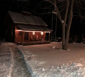 A wooden cabin with a porch, snow-covered ground, and a tree in front.