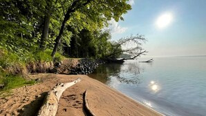 A sandy beach with a log and footprints, trees, and calm water. - Cozy Cabin Overlooking Chequamegon Bay in Ashland, Wisconsin (Ashland)