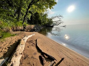 A sandy beach with a log and footprints, trees, and calm water. - Cozy Cabin Overlooking Chequamegon Bay in Ashland, Wisconsin (Ashland)