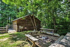 Una cabaña rústica con una terraza de madera, mesas de picnic y un hoyo para fogatas en un área boscosa.