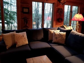 A cozy cabin interior with a sectional sofa, a wooden coffee table, and large windows offering a view of a forest. - Beautiful Log Cabin with Jacuzzi Bathtub in the heart of Adirondack State Park (Saranac Lake)