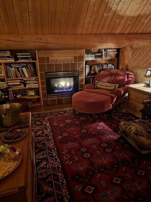 Une pièce chaleureuse avec une cheminée, un fauteuil rouge, une bibliothèque en bois et un tapis à motifs.