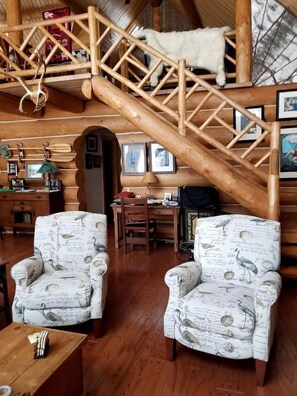 L’intérieur d’un chalet accueillant, avec un escalier en bois, deux fauteuils à motifs, un bureau et une lampe suspendue.