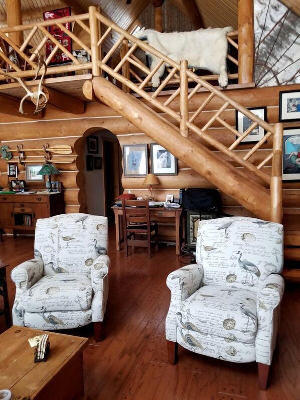 A cozy cabin interior with a wooden staircase, two patterned armchairs, a desk, and a hanging lamp. - Beautiful Log Cabin with Jacuzzi Bathtub in the heart of Adirondack State Park (Saranac Lake)