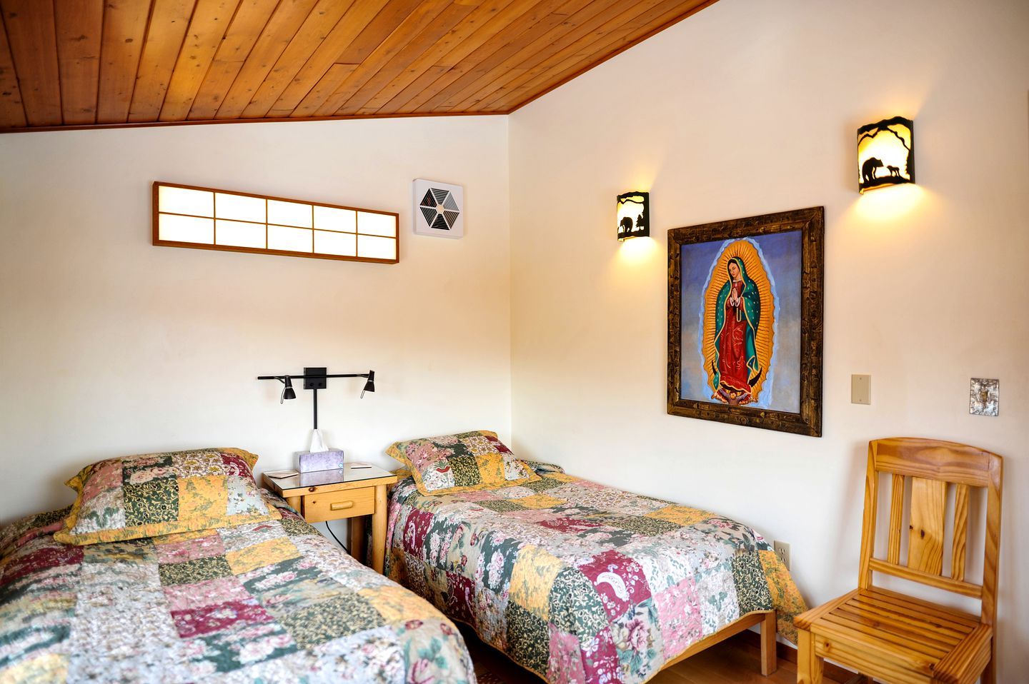 Quarto com duas camas, uma cadeira de madeira, uma mesinha de cabeceira, um respiradouro na parede e um quadro emoldurado.