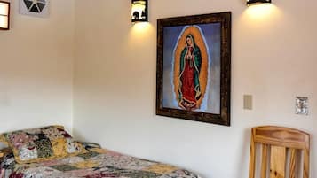 A room with a wooden ceiling, a bed with a floral bedspread, a wooden chair, and a framed religious painting.