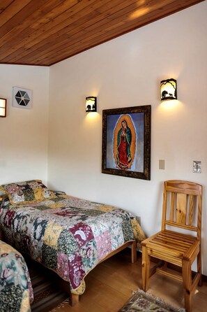 A room with a wooden ceiling, a bed with a floral bedspread, a wooden chair, and a framed religious painting.