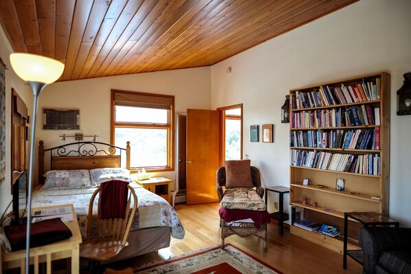A cozy room with a wooden ceiling, a bed with a floral bedspread, a chair with a cushion, a bookshelf filled with books, and a window letting in natural light. - Peaceful Luxury Vacation Rental near Gila National Forest in Silver City, New Mexico (Silver City)