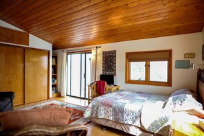 A bedroom with a wooden ceiling, a bed with a floral bedspread, a desk with a chair, a bookshelf, and a sliding glass door leading to a balcony. - Peaceful Luxury Vacation Rental near Gila National Forest in Silver City, New Mexico (Silver City)