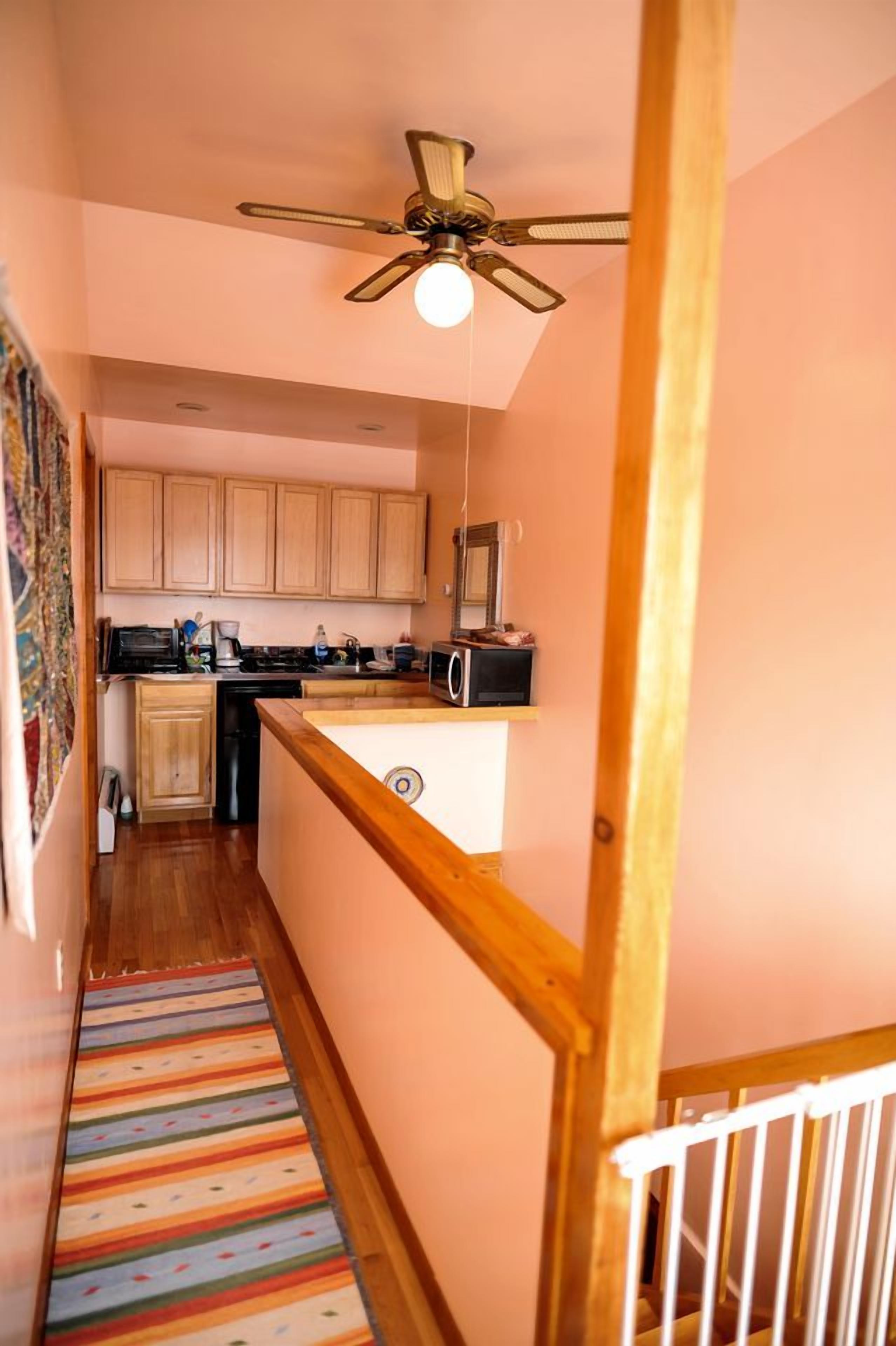 A narrow hallway with a wooden staircase, a ceiling fan, and a kitchen area with cabinets and appliances.