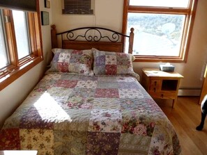 A bed with a floral quilt, a wooden headboard, a nightstand with a lamp, and a window with a view of a landscape. - Peaceful Luxury Vacation Rental near Gila National Forest in Silver City, New Mexico (Silver City)