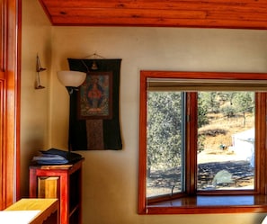A room with a wooden ceiling, a window with a view of a landscape, a wall-mounted lamp, and a small table with a book.
