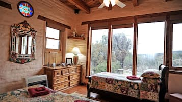 Una habitación rústica con vigas de madera, un ventanal con vistas, una cama con ropa de cama floral, un aparador y un espejo con marco decorativo.