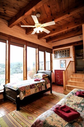 A wooden cabin bedroom with a bed, a bench, a staircase, and a view through large windows.