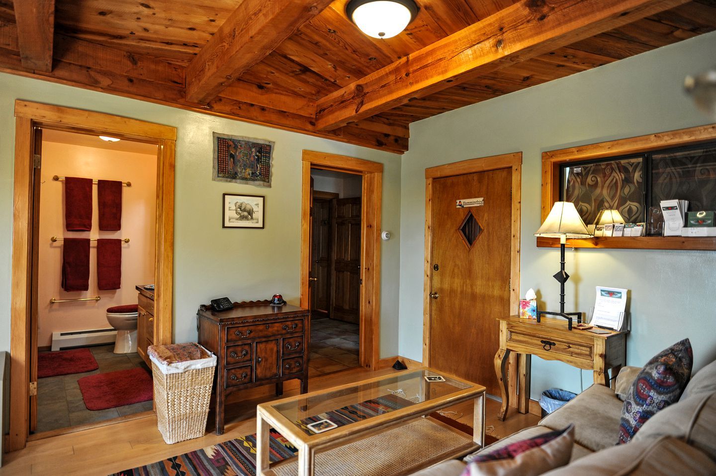 Un plafond en bois, une porte en bois, une table basse en verre, un canapé et un petit bureau en bois avec une lampe.