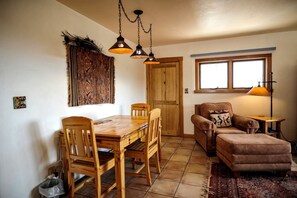 A dining area with a wooden table and chairs, a patterned rug, a sofa, and a window with a view.