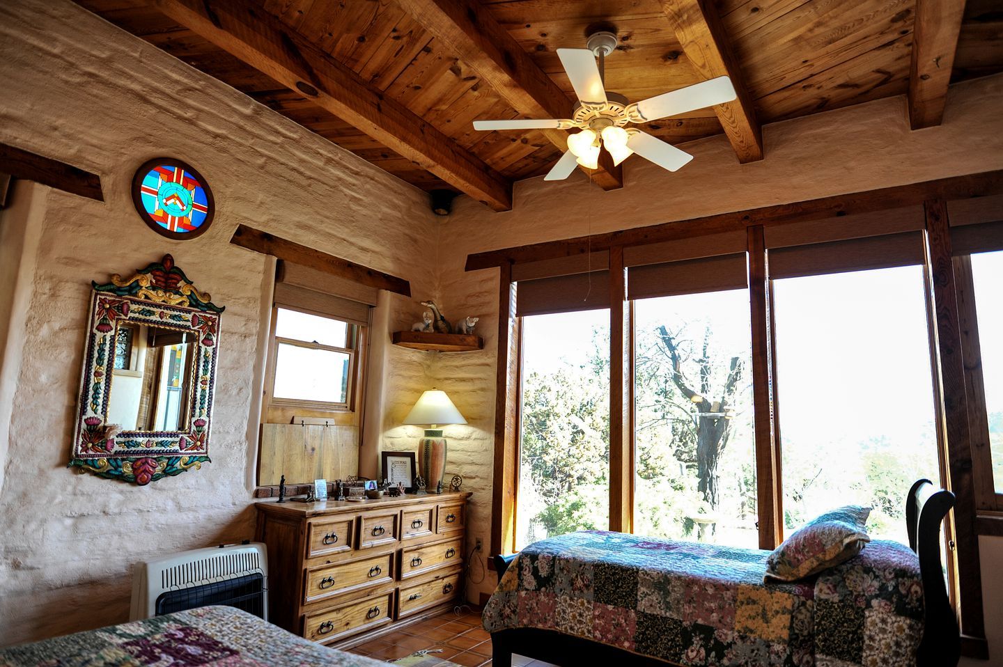 A rustic room with wooden beams, a ceiling fan, a large window with a view of trees, a dresser, and a bed with a quilt.