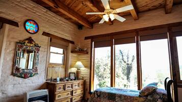 A rustic room with wooden beams, a ceiling fan, a large window with a view of trees, a dresser, and a bed with a quilt.