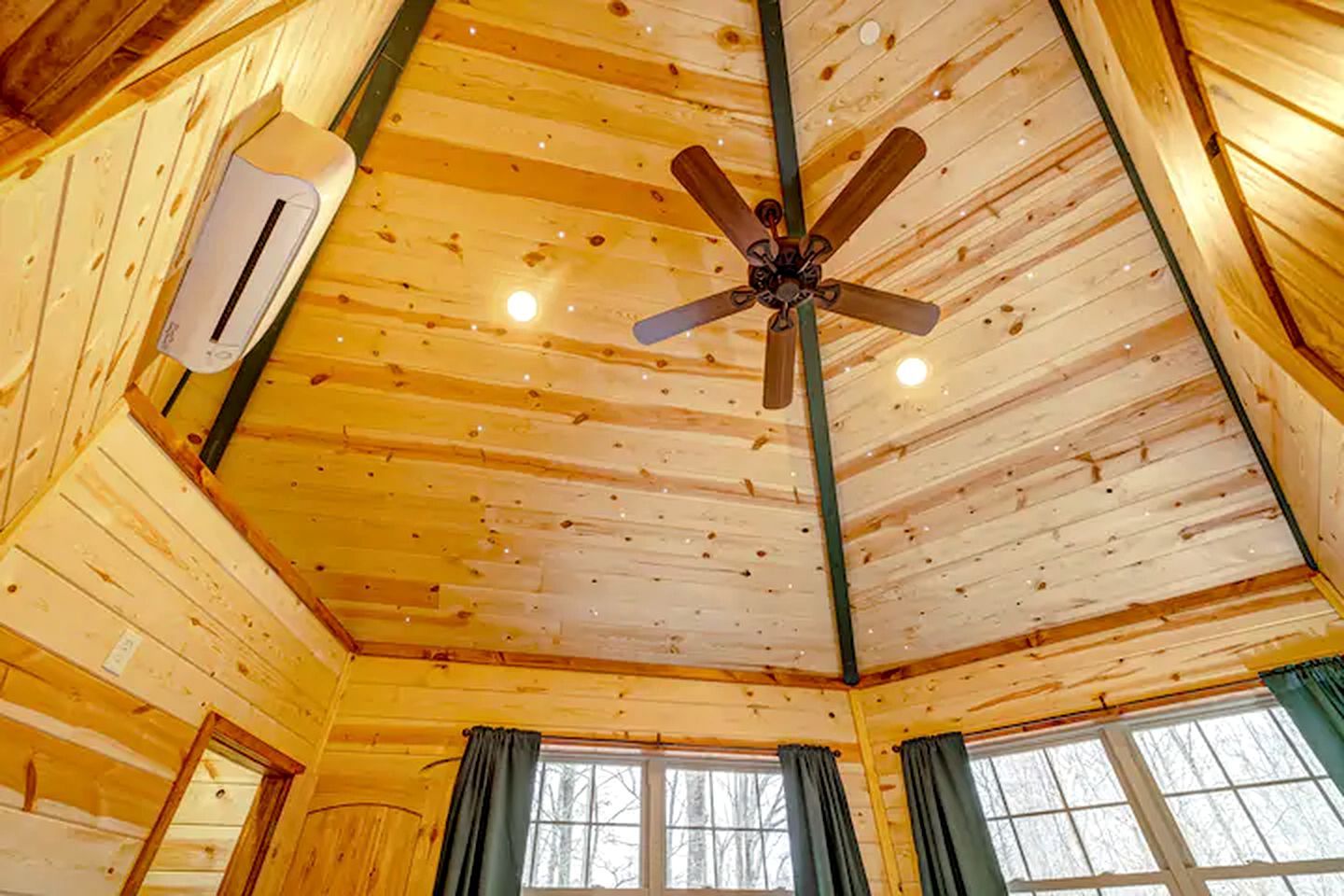 A cabin interior with a wooden ceiling, a ceiling fan, an air conditioning unit, and windows with curtains.