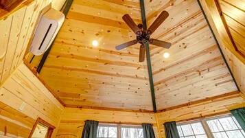 A cabin interior with a wooden ceiling, a ceiling fan, an air conditioning unit, and windows with curtains.
