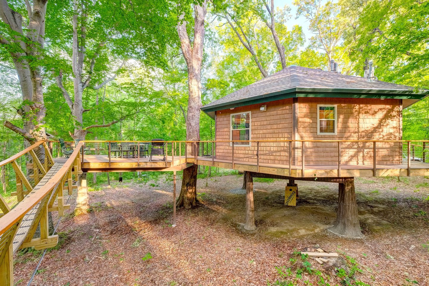 Una cabaña de madera sobre pilotes en un bosque, con una terraza de madera y un puente.