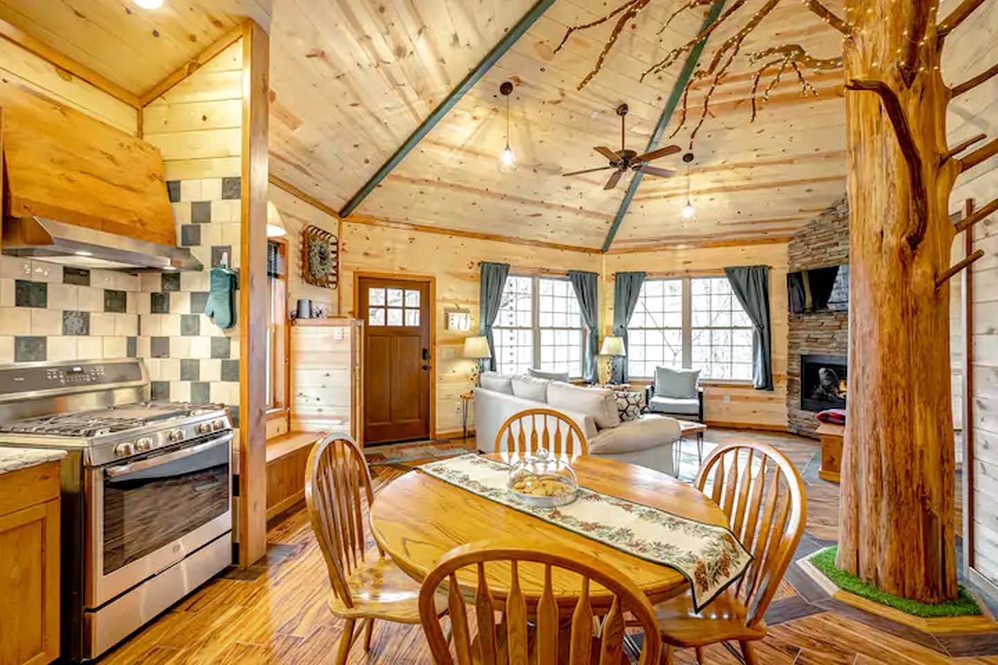 A wooden cabin interior with a kitchen, dining area, and living room, featuring a fireplace and large windows.