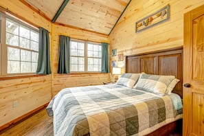 Une chambre dans une cabane en bois, avec un lit, deux fenêtres avec des rideaux, un plafond en bois et une porte.