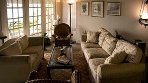 Un salon avec un canapé, un fauteuil et une table basse. De grandes fenêtres, une lampe et des tableaux encadrés ornent le mur.