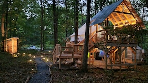A rustic wooden cabin with a balcony, surrounded by trees and a gravel path.