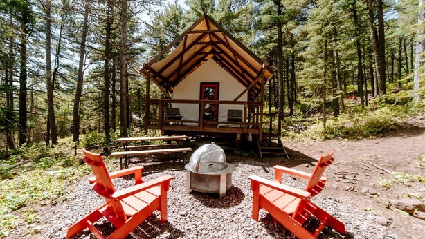 A cabin with a wooden deck, two red chairs, a picnic table, and a grill. - Glamping Safari Tent for Romantic Escapes to the Minnesota Forest (Beaver Bay)