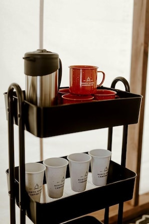 A black metal cart with a coffee pot and red cups on the top shelf, and white cups on the bottom shelf.