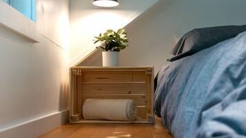A small, cozy room with a wooden floor, a bed with a blue blanket, a small wooden nightstand with a plant, and a wall-mounted light fixture.