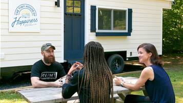 Three people seated at an outdoor wooden table with a white mobile home in the background.