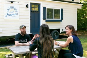 Three people seated at an outdoor wooden table with a white mobile home in the background.