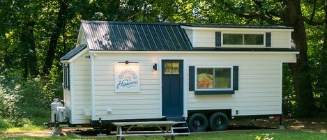 A mobile home with a small deck, a sign reading 'The Tiny House of Happiness,' and a window with blinds.