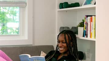 A person sitting on a grey sofa reading a book in a room with a white wall, a window with blinds, and a bookshelf with books and plants.