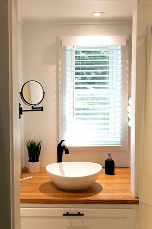 A bathroom with a white sink, a round mirror, a potted plant, and a window with blinds.