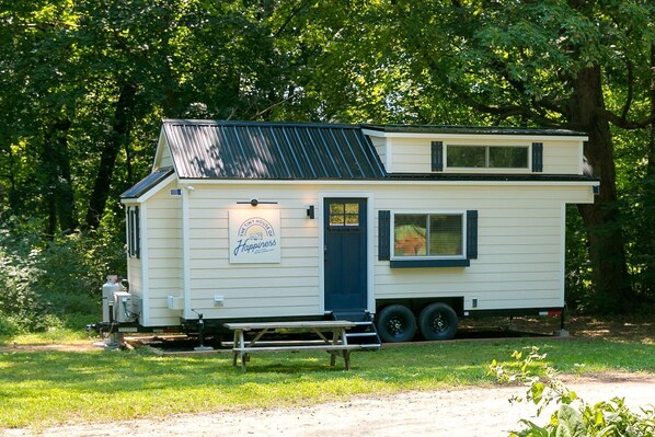 A mobile home with a small deck, a sign reading 'The Tiny House of Happiness,' and a window with blinds.