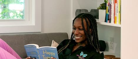 A person sitting on a grey sofa reading a book in a room with a white wall, a window with blinds, and a bookshelf with books and plants.