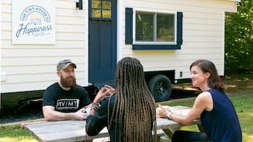 Three people seated at an outdoor wooden table with a white mobile home in the background.