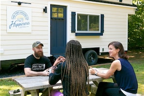 Trois personnes sont assises à une table en bois à l’extérieur, avec une caravane blanche en arrière-plan.