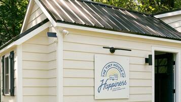 A small, single-story building with a metal roof and a sign that reads 'The Tiny House of Happiness: Less Stuff, More Connection.'