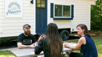 Three people seated at an outdoor wooden table with a white mobile home in the background.