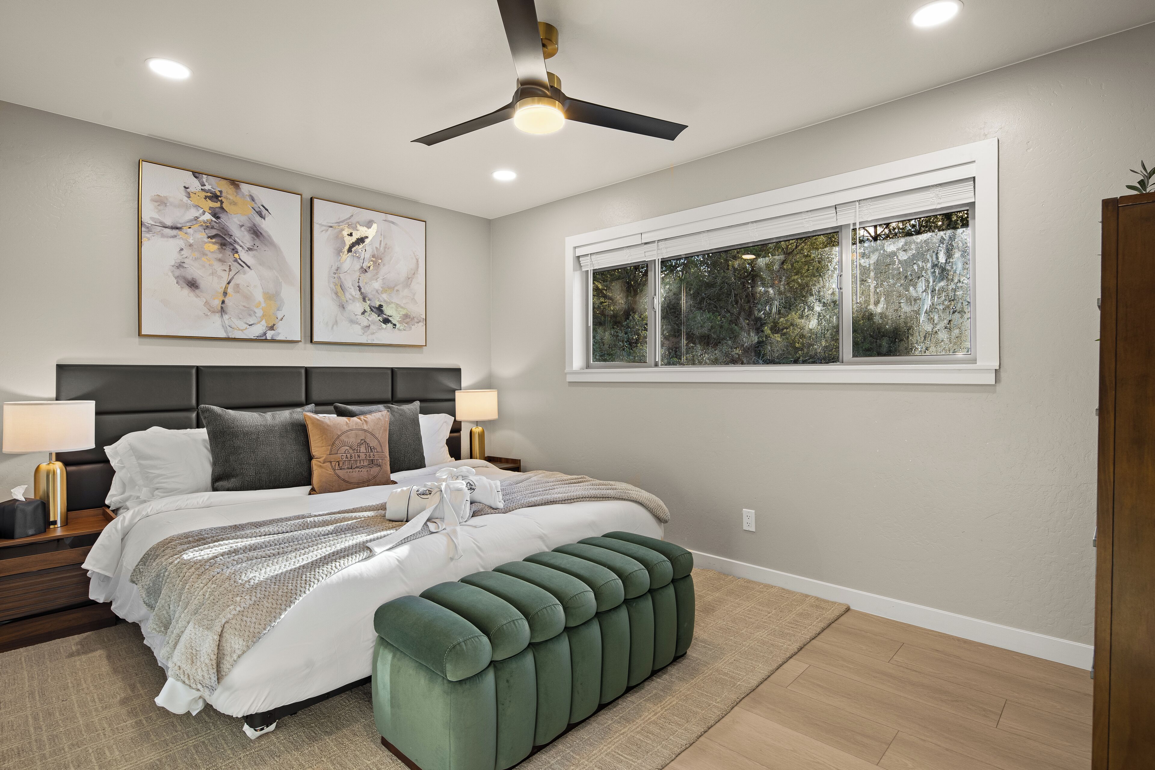 A bedroom with a bed, nightstands, a bench, a ceiling fan, and a window with a view of trees.