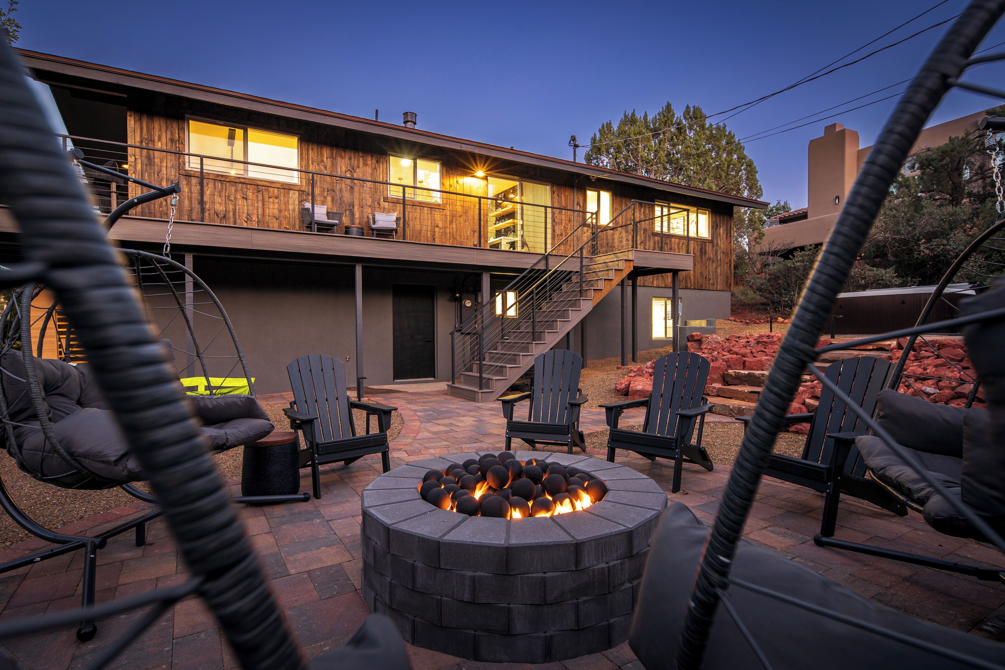 A modern outdoor patio with a circular fire pit, outdoor seating, and a staircase leading to an upper level of a wooden house.