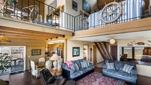 A spacious living area with a high ceiling, wooden beams, a balcony with a ship's wheel, and a dining area with a table and chairs. - Fantastic Beach View! Oregon Escape w/ Game Room (Seaside)